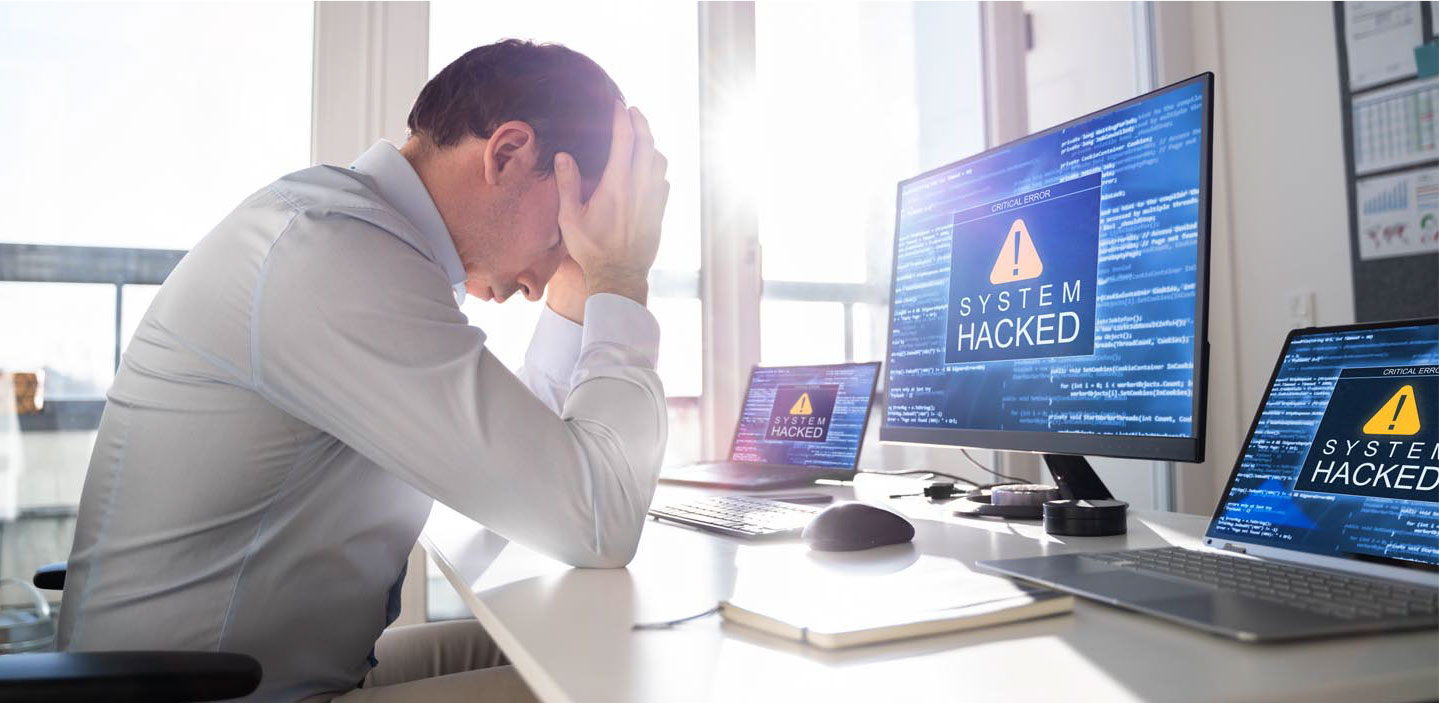 Show thumbnail preview Ein Mann sitzt mit dem Kopf in den Händen an einem Schreibtisch, umgeben von Computerbildschirmen, auf denen die Warnung „System gehackt“ angezeigt wird.